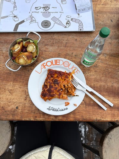 An overhead view captures a meal served on a rustic wooden table at La Folie Douce, Méribel Mottaret, 73550 Les Allues, France. The scene is outdoors, indicated by snow visible on the ground beneath the table and a wooden stool.
On the table, a white ceramic plate, branded "LA FOLIE DOUCE" and "La Petite Cuisine," holds a partially consumed slice of baked pasta, resembling lasagna, with a browned, crispy top. A silver fork and knife rest on the plate's right side. To the upper left of the plate, a small, silver-toned metal pot with handles contains four individual servings of what appear to be toasted bread rolls topped with round slices of goat cheese, garnished with green leaves and red cranberries. A clear plastic bottle of Badoit mineral water with a green cap stands to the right of the plate.
Above the food items, a laminated placemat or menu is visible, featuring black and white illustrations of hands holding wine glasses and bottles, with the text "You survived Folie Douce ?" and "Mon Chéri." A partially obscured white receipt, confirming "LA FOLIE DOUCE" and the Méribel Mottaret, Les Allues location, is tucked under the placemat. The lower body of an individual, wearing black pants and a light-colored quilted jacket, is visible seated at the table, suggesting a first-person perspective. The wooden table surface is worn with visible grain and slight indentations.