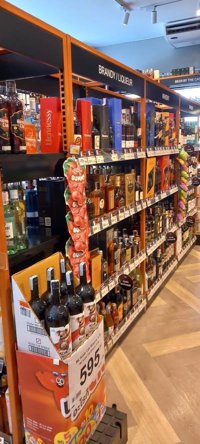 The image displays an aisle in a retail store, likely a liquor store or supermarket, filled with various alcoholic beverages. The shelves are a combination of orange and black, vertically aligned and stocked with numerous bottles. Overhead signs categorize the products, with "BRANDY / LIQUEUR" clearly visible, and "WHISKY" and "BEER BY THE CAS" partially seen further down the aisle.

Specific brands of spirits are identifiable, including "Hennessy VSOP", "Martell" (partially visible), and "Havana Club". A prominent display box contains "KOALA" branded wines, with labels such as "KOALA CAB RESERVE" and "KOALA ROSSINA". Many small price tags are attached to the shelves, showing various numbers like "78", "32", "549", "699", and a larger tag with "595". Promotional text such as "Just arrived!" and "GUMMY" are also present. The KOALA wine box features a cartoon character and Thai text including "อะไรก็" and "ลด" (meaning 'discount' or 'sale'), along with dates "14 Mar 2025 - 30 Apr 2026" which likely refer to a promotional period.

The setting is indoors, with an air conditioning unit and ceiling lights visible. The floor has a light-colored wooden herringbone pattern. The scene depicts a typical retail environment, with products displayed for sale. Given the presence of Thai text and the provided location context, the store is situated in Moo 1, Thailand.