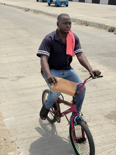 A male subject, estimated to be in his 20s to 30s, with short dark hair and dark skin, is depicted riding a bicycle on a concrete road in Jakande, Nigeria. He is wearing a dark blue short-sleeved collared shirt with two white horizontal stripes on each sleeve, light blue denim jeans with visible wear, and dark open-toed sandals. A red towel is draped over his left shoulder and neck. He holds a rectangular, light brown, flat object in his left hand; a silver ring is visible on his left pinky finger. His right hand grips the right handlebar of the bicycle. The subject's gaze is directed towards the upper right of the frame.
The bicycle is a small, BMX-style model, predominantly painted a faded and chipped magenta/pink. Its wheel rims are a distinct light green or aqua color, and a light blue plastic piece is attached to the front fork area.
The environment consists of a wide, light-colored concrete road. To the left side of the frame, in the background, a raised concrete curb separates the current road section from another paved area where two light blue, three-wheeled utility vehicles are visible in the distance, traveling away from the viewer. Scattered small debris is present on the road surface. Strong shadows are cast on the ground, indicating direct overhead sunlight.