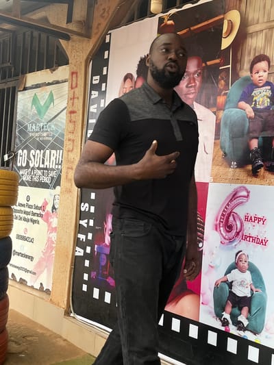 This media file shows a man posing in front of large posters. The man is wearing a black polo shirt and dark pants. He has a beard and is looking towards the camera. Behind him, there are several posters. One poster depicts a child on a green chair with "HAPPY BIRTHDAY" written above. Another poster features the text "DAVID SYFA". Further down, a poster advertises "MARTECK ENERGY LIMITED" with the slogan "GO SOLAR!! MAKE IT A POINT TO SAVE MORE THIS PERIOD." The advertisement also includes contact information, an address in Dei Dei, Abuja, Nigeria, a phone number, and an email address. The setting appears to be outdoors, possibly a street or an open-air market in Sabonjibi, Nigeria, given the context provided. The overall impression is a casual setting with promotional materials displayed.