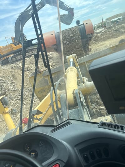 The image shows a view from inside the cab of a Volvo construction vehicle, likely a wheel loader, at an apparent construction or excavation site. The Volvo excavator is also visible outside, on what appears to be gravel or rocky terrain. Other construction equipment, including a cylindrical orange piece, is visible in the background, along with what may be scaffolding or framework. Based on the provided information the location is at an apparent construction site in the city of Elenino, Bulgaria. However, there are no people visible in the scene.