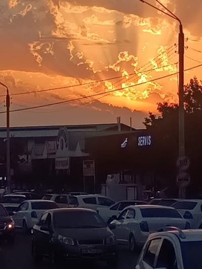 In Yalangach, Uzbekistan, a traffic jam unfolds at dusk under a dramatic sky. The sky is filled with luminous, orange and yellow clouds, illuminated by the setting sun, with visible sunbeams piercing through. In the mid-ground, a dark, modern building features a prominent sign that reads "SERVIS" in white illuminated letters, accompanied by a stylized white logo. Several cars are visible in the foreground, forming a queue on a road. Streetlights and utility wires crisscross the scene, adding to the urban setting. The overall mood is one of a busy city street experiencing a transition from day to night, characterized by traffic, commerce, and a striking natural sky.