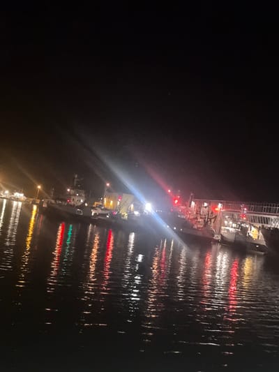 The image depicts a nighttime scene at a harbor in Husøy, Norway. The upper half of the frame shows a dark, clear night sky. The lower half displays a body of water, likely a bay or inlet, exhibiting numerous elongated reflections of artificial light sources. Several vessels are docked along a visible shoreline or pier extending from the left to the right side of the image.
On the left, a portion of what appears to be a dock or industrial area is visible with yellow-orange lighting. In the mid-ground, at least two boats or ships are moored. One larger vessel is prominent towards the right, featuring multiple decks, structures, and what appear to be fishing or cargo handling equipment. A smaller vessel is positioned to its left.
The scene is illuminated by various artificial lights, including white, yellow, orange, green, and red sources. A bright, white light source emits a strong glare from the mid-right side of the image, extending a prominent streak across the water. Several distinct red lights are visible on the vessels and shore structures, casting corresponding red reflections. Yellow-orange lights are visible further left, also reflecting in the water. No individuals are discernible within the frame. The water surface shows ripple patterns, distorted by the light reflections.