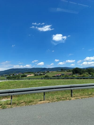 The image shows a wide, horizontal shot of a rural landscape under a clear blue sky with scattered white clouds. In the foreground, a section of grey asphalt road is visible, followed by a metallic guardrail separating it from a grassy verge and a field. The field is green and appears to be well-maintained, with some dry patches and scattered yellow flowers.
Beyond the field, rolling green hills rise, dotted with trees and several residential buildings with red-tiled roofs. Some of these buildings are clustered together, forming small hamlets or villages. The architecture suggests a European setting, likely a small town or rural area. The hills in the background are covered in lush green vegetation, and the horizon is marked by a range of more distant, darker green mountains. The overall impression is one of a peaceful, sunny day in the countryside.
While the image depicts a general rural landscape, the prompt asks for a specific location in Niederwinkling, Germany. Without additional information or visual cues that are specific to Niederwinkling, it is impossible to definitively confirm or deny the location. However, if this scene is indeed in Niederwinkling, it suggests that the area features rolling hills, agricultural fields, scattered settlements with traditional European architecture, and is bordered by mountainous terrain. The presence of a road with a guardrail indicates it could be near a main thoroughfare or a scenic route. There are no people or animals visible in the image.