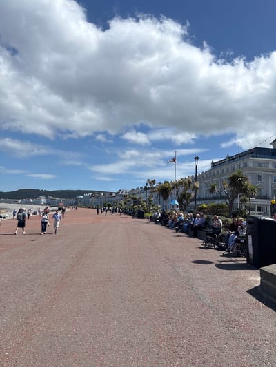The image presents a view of the seafront promenade in Llandudno, United Kingdom, under a partly cloudy sky. A wide walkway, textured in a reddish-brown, dominates the foreground and extends into the distance. Along the edge of the walkway closest to the buildings, a series of benches are occupied by people seated and facing the sea. Some individuals appear to be in wheelchairs. The buildings lining the promenade are light-colored, multi-story structures featuring architectural details like balconies and awnings. Palm trees are interspersed along the building front. A flag is visible, flying from a pole in front of the buildings. In the distance, the coastline stretches with buildings and a backdrop of a wooded hill or small mountain. Some people can be seen walking and relaxing on the beach below the promenade.
