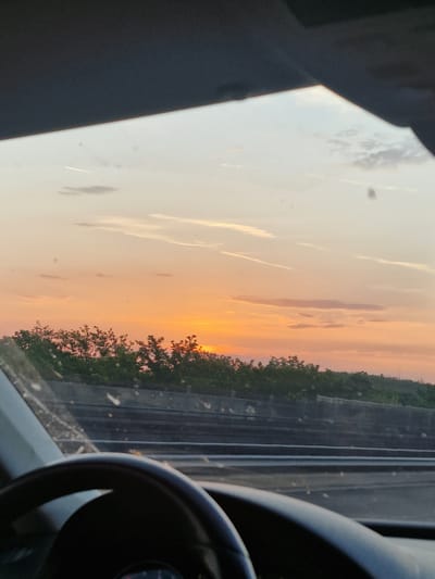 The image captures a view from inside a car, looking through the windshield at a sunset over Balatonszemes, Hungary. The sky is a gradient of warm orange and soft yellow, with wispy clouds scattered across it. A thin contrail from an airplane is visible in the upper left portion of the sky. Below the sky, a line of dense green trees creates a natural barrier. In the foreground, the steering wheel of the car is partially visible, indicating the perspective of the driver. The windshield has several small white specks, likely dust or water droplets. The scene suggests a peaceful drive during golden hour.
