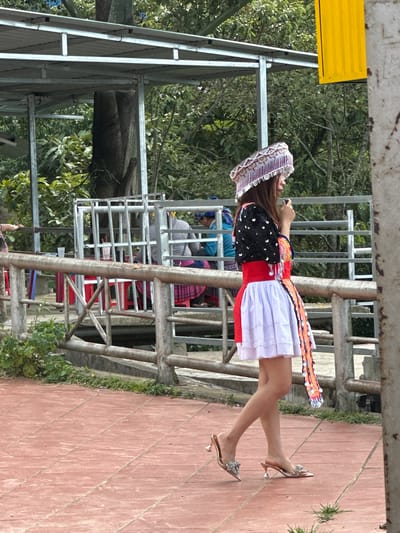 In Tu Le, Vietnam, a young woman, facing left in profile, walks across a reddish-brown tiled pavement. She is dressed in traditional attire, featuring an elaborate patterned silver-and-white headpiece with dangling ornaments, a black short-sleeved top with white butterfly-like embellishments, a wide red waist belt, and a white pleated mini-skirt with a red panel on the left side. A long, colorful, beaded or sequined sash hangs from her left hip. She wears low, pointed-toe, high-heeled mules with sparkling embellishments on the toe strap. Her right hand holds a small dark object, possibly a phone or microphone, near her face.
Behind her, a concrete railing with metal posts and horizontal bars separates the walkway from an area below. Further back, an open-air structure with a corrugated metal roof supported by metal framework provides shelter. Beneath this structure, several individuals are visible, seated at tables with red plastic chairs. One person on the far left wears a blue top, and another to the right wears a grey top and a patterned pink skirt. Lush green trees and foliage dominate the background, with a large tree trunk visible centrally. On the far right, a partial view of a bright yellow rectangular sign or banner is visible next to a grey pillar. Green vegetation sprouts intermittently along the edge of the pavement and railing.