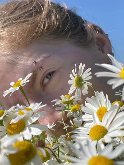 A close-up shot captures a person's face partially obscured by a cluster of white daisies with yellow centers. The person has fair skin with a scattering of freckles, and their left eye, a shade of green, is visible. Two silver eyebrow piercings are present. Their hair, a light blonde color, is messy and windswept. Multiple silver piercings adorn their ear, which is also visible. The background is a clear, bright blue sky. The overall impression is one of natural light and a focus on the textures of skin and petals, with an implied outdoor setting. The image does not contain any information about the city of Сям-Можга, Russia.