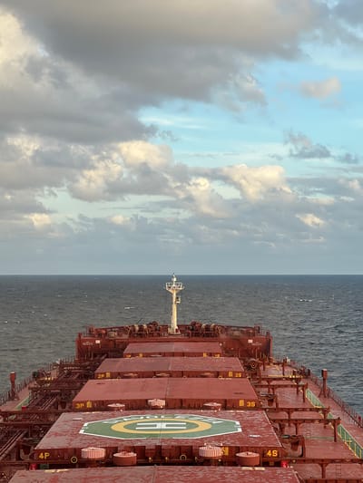 The image shows a perspective from the deck of a large cargo ship sailing on the ocean. The ship's deck is painted a reddish-brown and is covered with numerous hatches and piping. In the foreground, a helipad is visible on one of the hatches, marked with a green circle and yellow lines. The ship's mast with navigational lights stands tall in the middle of the frame. The sea is a dark, choppy blue, stretching out to the horizon. The sky above is filled with a mix of grey and white clouds, with patches of pale blue sky peeking through. The lighting suggests it is either early morning or late afternoon. The location in the city of Unknown, Papua New Guinea is not determinable from the image.
