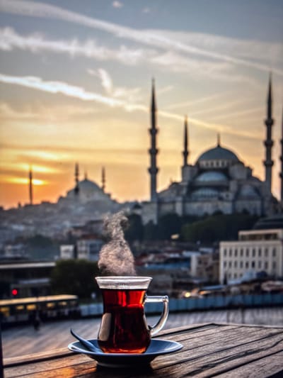 A cup of hot tea sits on a wooden table in the foreground, with steam rising from it. In the background, a city skyline is visible, dominated by a large mosque with multiple minarets. The sky is overcast with streaks of clouds and a warm glow from the setting sun. The city appears to be located in a coastal region, with hints of water and buildings along the shoreline. The overall impression is one of tranquility and a moment of relaxation. It is impossible to confirm the location as Pastavy, Belarus, based on the visual evidence.