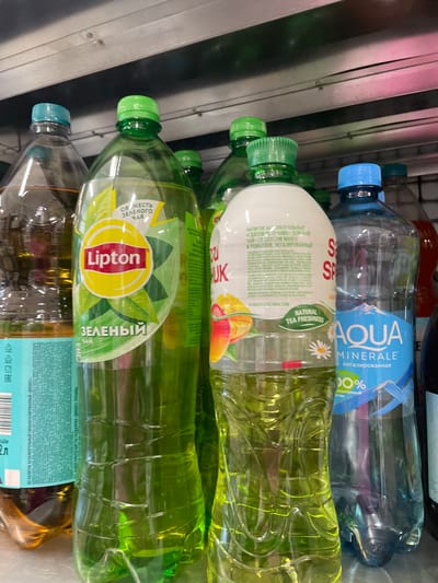 In Ladozhskii Trudposelok, Russia, a shelf display features an assortment of beverages. Dominating the foreground are several large plastic bottles of Lipton green tea, their vibrant green liquid and prominent yellow and red logos clearly visible. To the right, a bottle of Aqua Minerale water with a blue label showcasing mountain imagery and the percentage "100%" stands out. To the left of the Lipton bottles, partially obscured, is another beverage bottle with a teal-colored label. The bottles are arranged in a staggered fashion, suggesting a retail environment such as a grocery store or convenience store. The lighting in the scene appears to be overhead store lighting.