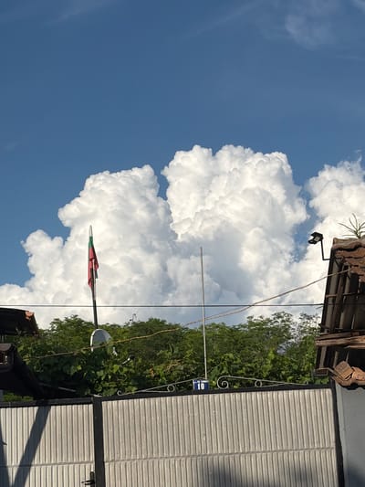 The image depicts a bright, sunny day with a clear blue sky and large, fluffy white clouds. In the foreground, a corrugated metal gate is visible, casting a long shadow on its left side. Above the gate, a row of green trees and foliage forms a natural barrier. A white satellite dish is mounted on a pole amidst the greenery.
To the left of the foliage, a flagpole stands tall, bearing a Bulgarian flag with its white, green, and red horizontal stripes. Further to the right, a portion of a tiled roof is visible, with a modern black outdoor light fixture attached to its side. A street sign with the number "10" and some text in Bulgarian (likely the street name) is mounted on a decorative metal fence above the gate. The overall scene suggests a residential area in a town or village, with the Bulgarian flag and the street sign providing clues to the location. The setting appears to be the city of Kozlovets, Bulgaria, based on the presence of the Bulgarian flag and the visual aesthetic of the buildings and natural surroundings, which are consistent with typical Bulgarian rural or semi-urban landscapes.