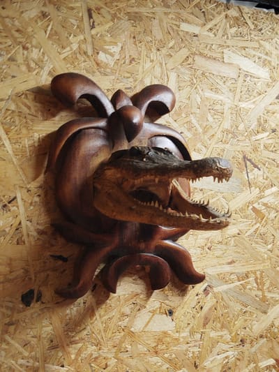 A wooden alligator head with an open mouth is mounted on a wooden Fleur de Lis plaque, which is attached to a wooden panel wall. The location is Боровково, Russia.