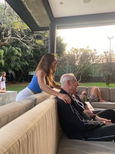 Outdoors, a woman in a blue top and white pants attends to a seated older man in a black jacket and pants with white stripes. The man rests on a light-colored outdoor sofa. Another person's legs are visible in the background, also seated on the sofa. Trees and foliage fill the background, with a glimpse of a white tennis court fence. An additional figure, smaller in scale, stands below the woman and the man.