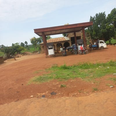 In Dorowa, Nigeria, a rural gas station is depicted under a partly cloudy sky. The station features a covered canopy with a corrugated metal roof, supported by red-brown pillars. A white fuel pump with a black screen stands under the canopy. Several people are present, including two individuals on a red motorcycle, one wearing a white helmet and a dark jacket with the number "09" on the back, and another in a red hooded sweatshirt. To the right, a white truck with a blue cargo bed is parked, with several people gathered around it. A ladder is leaning against the building behind the truck. The ground is covered in red-brown dirt and patches of green grass. In the background, trees and vegetation are visible, with some farm fields in the distance. The overall scene suggests a functional, albeit rustic, stop for fuel and transport in a rural Nigerian setting.