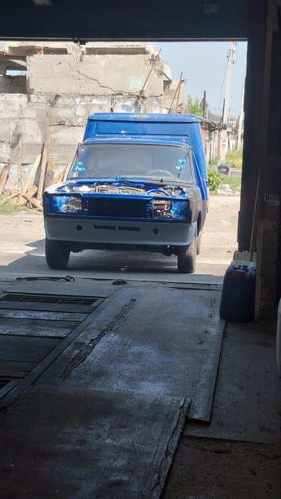 The image depicts a blue car, possibly undergoing repairs or customization, positioned outside an open garage or workshop. The car's hood is open, revealing the engine compartment. The immediate foreground shows a concrete floor with metal plates and a drainage grate. In the background, a dilapidated building structure and construction materials are visible, suggesting an urban or industrial environment. The lighting indicates it is daytime, with shadows cast across the scene. There are no people visible in the image. The location is identified as Belovo, Russia.