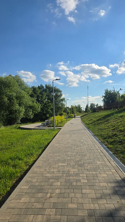 This media file depicts an outdoor scene in Razvilka, Russia. The primary subjects are a paved walkway, a grassy slope, trees, and a bright blue sky with scattered white clouds. The walkway, made of interlocking rectangular pavers, stretches into the distance, bordered by a strip of green grass and a metal grate along the edge of the grassy slope. Several lampposts with modern designs are visible along the walkway, suggesting it's a public space like a park or a promenade.
In the mid-ground, a single person is walking on the path, appearing as a small figure in the distance. There are also benches visible, hinting at a place for rest and relaxation. The scene is bathed in sunlight, indicating it is daytime. The clear blue sky and fluffy clouds suggest fair weather. The lush green grass and full foliage on the trees point towards a warm season, likely spring or summer. There are no visible text elements in the image. The overall impression is one of a peaceful, well-maintained outdoor environment.