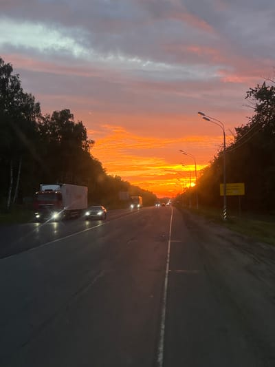 The image depicts a road scene during a vibrant sunset, likely in or near the city of Gazoprovodsk, Russia. The sky is dominated by intense shades of orange and yellow, with streaks of pink and purple, indicating a dramatic twilight. Tall, dark trees line the sides of the road, creating a silhouette against the bright sky. Several vehicles are visible on the asphalt road. On the left, a white truck with a red cab is prominent, its headlights illuminating the wet road surface. To the right of the truck, a silver car is also driving, its headlights on. Further down the road, other vehicles are present, their lights visible in the distance. Streetlights with multiple lamps are mounted on poles along the right side of the road, their lights also on. A yellow sign is affixed to a pole on the right side of the road, though its text is illegible. The road itself appears to be a multi-lane highway, with a white dashed line marking the lanes. The overall impression is one of a journey along a highway at dusk, with the natural beauty of the sunset contrasting with the man-made elements of the road and vehicles.