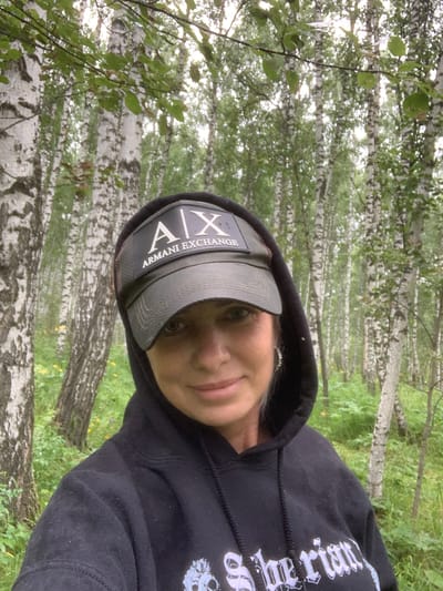 A woman wearing a black hoodie and an Armani Exchange baseball cap is taking a selfie in a birch forest. She has blonde hair and is wearing earrings. The forest is lush with green foliage and tall birch trees with white bark. The ground is covered in grass and some yellow wildflowers. The sky is overcast, suggesting a cloudy day. The location is identified as the city of Uyar, Russia.