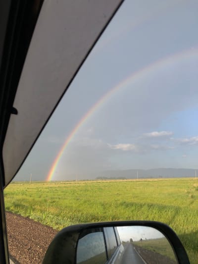 The image captures a view from inside a vehicle, looking out towards a rural landscape. A prominent rainbow arcs across a pale blue and grey sky. In the foreground, the edge of the car's window frame is visible on the left. The road, covered in gravel, runs along the left side of the frame. A vast, green grassy field stretches out towards a distant line of hazy mountains. The side-view mirror of the car reflects a paved road with a car driving ahead, and some vegetation on the side. The overall scene suggests a drive through the countryside, possibly after or during rainfall, given the presence of the rainbow and the slightly wet appearance of the road in the mirror. The location is identified as the city of Райков, Russia.
