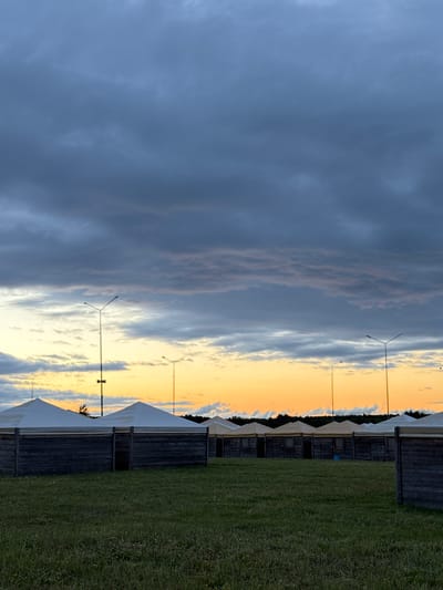 The image depicts a row of temporary, tent-like structures with wooden bases, set up on a grassy field. The structures have white or tan peaked roofs. In the background, several lampposts are visible, and a line of trees forms the horizon. The sky is filled with dramatic clouds, transitioning from dark grey at the top to bright orange and yellow hues closer to the horizon, suggesting either sunrise or sunset. The scene is located in the city of Bolgar, Russia. There are no people visible in the image.