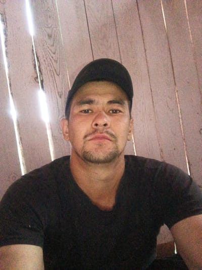 A man wearing a black t-shirt and a black baseball cap is pictured in a close-up, waist-up shot. He is positioned in front of a wooden wall with vertical planks. The man has dark hair and is looking directly at the camera. His facial features include a mustache and a goatee. The lighting suggests the photo was taken outdoors or near an opening, with bright light filtering in from the left side of the frame. The image does not provide any specific details about the location within the city of Islambaevo, Russia, beyond the implied setting of a wooden structure.