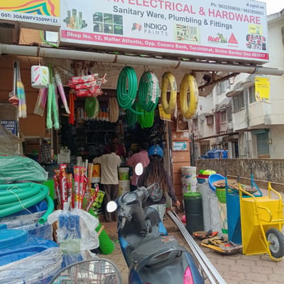 The image captures the exterior of "AR Electrical & Hardware," a shop in Siolim Bardez, Goa, India (postal code 403517), as indicated by the sign. The shop sells sanitary ware, plumbing, and hardware. The storefront is cluttered with merchandise, including hoses, cleaning supplies, paint buckets, and PVC pipes. A blue scooter is parked in front, partially obstructing the view. Several people are visible, including shop staff and a customer with long dreadlocks wearing a helmet. The sign also lists two phone numbers, a GST number, and advertises Indigo Paints and Roff products. A yellow wheelbarrow is also visible to the right of the image. Across the street, a man is seen on the balcony. The overall scene is a busy, typical small business environment in India.