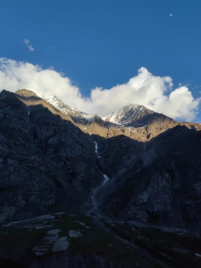 This media file depicts a breathtaking mountain landscape in Kead, India. The scene is dominated by imposing, rocky mountain peaks, some of which are capped with snow, suggesting a high-altitude, possibly alpine, environment. The mountains are dramatically lit by the sun, with stark shadows contrasting with sunlit areas, indicating it is either early morning or late afternoon. Several fluffy white clouds are scattered across a vibrant blue sky. A small crescent moon is visible in the upper right corner of the frame, further reinforcing the time of day.
In the foreground, the terrain appears to be terraced, with visible patches of greenery and what look like agricultural plots. A small, winding stream or river is visible flowing down through the mountainside, adding a sense of natural beauty and life to the rugged landscape. The overall atmosphere is one of serene grandeur and natural majesty. There is no visible text in the media file.