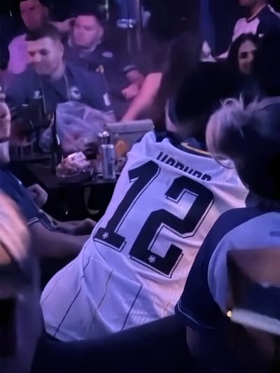 The image shows a group of people gathered indoors, possibly at a bar or restaurant, in Travnik, Bosnia and Herzegovina. The lighting is dim and appears to be colored, with prominent purple hues. In the foreground, a person wearing a white sports jersey with the number "12" and the word "HADŽIBEGIC" printed in black across the upper back is partially visible. Other individuals are seen in the background, some with their faces obscured. Objects on a table in the middle ground include a bottle, a glass, and what appears to be food. The overall atmosphere suggests a social gathering.