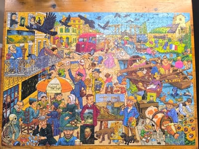 The image is a jigsaw puzzle depicting a whimsical, crowded scene set in the city of Uithoorn, Netherlands, with a prominent focus on artist Vincent van Gogh. The scene is a busy street and canal, populated by numerous caricatured figures engaged in various activities. In the foreground, a skeleton sits at a table outside a cafe, smoking a cigarette. Adjacent to this, a man with a beard and wearing a hat is depicted with a beer. A cafe with an umbrella labeled "Cafetaria Vincent van Gogh" is visible, with patrons seated at tables.
Further into the scene, a red van with the text "CASSE VERHUIZINGEN" is parked. People are shown interacting with each other, some walking, others riding bicycles or scooters. A canal runs through the center of the image, with several boats carrying passengers. One boat has a French flag.
Artist Vincent van Gogh is represented multiple times, including a painting of a bridge, and a man resembling him is shown painting sunflowers. Several sunflowers are also depicted in the foreground. Birds, including crows, are present in the sky and on buildings. The architecture suggests a European setting with buildings featuring yellow facades and tile roofs. The overall atmosphere is lively and humorous.