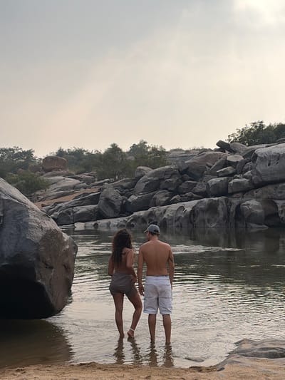 Two individuals stand waist-deep in a shallow river, viewed from behind, in a rocky landscape identified as Hale Bandi Harlapura, India. The person on the left, with long dark curly hair, wears a light-colored short skirt or shorts and appears bare-chested or in a bra. The person on the right, a male, wears white shorts or swim trunks and a light-colored baseball cap, and is bare-chested. Their reflections are visible in the calm water. They are positioned near a sandy bank on the left foreground. The river is surrounded by numerous large, smooth, grey boulders and rock formations, some with distinct erosional features. Sparse green vegetation, including small trees and bushes, grows among the rocks on the distant banks. The sky above is overcast and light grey. A very large, rounded boulder occupies the immediate left foreground.