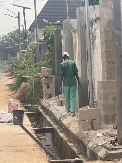 An outdoor scene shows a person wearing green work clothes and a white hat standing next to a constructed concrete structure with multiple recessed openings. To the side are construction materials: bricks, cement bags, and a bucket. Vegetation and street lamps line a road in the background, with a glimpse of houses visible beyond.