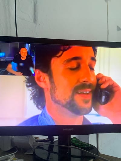 The image shows a television screen displaying a video with two distinct scenes. The primary scene, taking up most of the screen, shows a close-up, upside-down shot of a man's face. He has dark hair and a beard, and his mouth is slightly open as if speaking or reacting. A hand, also upside down, is visible near his head, holding a dark object, possibly a remote control or a phone receiver. The lighting on his face is warm and orange-toned.
Below this main scene, in the lower left corner of the screen, is a smaller, rectangular inset video. This inset also shows a man, right-side up, wearing a dark shirt. He is looking towards the camera with a neutral expression. The background of this inset appears to be a room with some blue and purple lighting effects.
On the right side of the television screen, part of the frame of the television itself is visible, along with some objects behind it. These include a power strip with cables and adapters, and a dark object that looks like a shoe or flip-flop. The Philips logo is visible on the lower right side of the television frame.
The overall impression is that this is a recording or broadcast being displayed on a TV. The orientation of the main subject suggests the video might have been filmed in a way that requires rotation for proper viewing. The setting appears to be indoors, likely a living room or entertainment area, given the presence of the television and its accessories. No specific time of day or weather conditions can be inferred. The emotions of the main subject in the primary scene are unclear due to the close-up and upside-down orientation, but the inset subject appears calm.