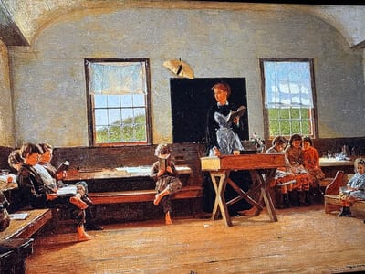 This image depicts a brightly lit interior of a historical classroom, characterized by light-colored textured walls and a wooden floor. Two large multi-pane windows with sheer white coverings illuminate the room from the left and right, revealing green foliage outside. An arched ceiling element is visible in the upper corners.

In the center-right, a female teacher stands behind a simple wooden lectern with an X-shaped base, holding an open book. She has auburn hair pulled back and wears a dark dress with a light-colored decorative bodice or apron. Behind her, a dark rectangular blackboard is mounted on the wall, above which a light-colored, possibly straw, fan or hat is pinned.

On the left side of the room, several male students are seated on long wooden benches at desks, engaged in reading. One student in the foreground of this group has bare feet resting on the floor. A lone student, also barefoot and wearing dark, loose clothing, sits on a bench in the mid-left foreground, intently reading an open book held close to his face.

On the right side of the room, younger students are seated. Three students are positioned at a desk to the immediate right of the teacher, while a young girl in a light-colored dress and dark boots sits on a separate wooden bench in the far right foreground, also engrossed in reading a book. A small arrangement of flowers in a vase is present on the teacher's lectern.

The image provides no identifiable details linking the depicted classroom or its contents to the city of Reus, Spain.