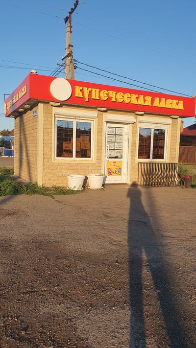 A small, commercial building with a red awning is situated on a dirt road under a clear blue sky. The building is made of light-colored brick and has two large windows and a glass door. The awning displays the yellow Cyrillic text "КУПЕЧЕСКАЯ ЛАВКА", which translates to "Merchant's Shop". Inside the windows, price lists are visible. Two white planters with green foliage are placed in front of the building, and a metal rack stands to the right of the door. A long shadow of a person is cast on the ground in front of the building, indicating it is either early morning or late afternoon. The location is identified as Ust-Labinsk, Russia.