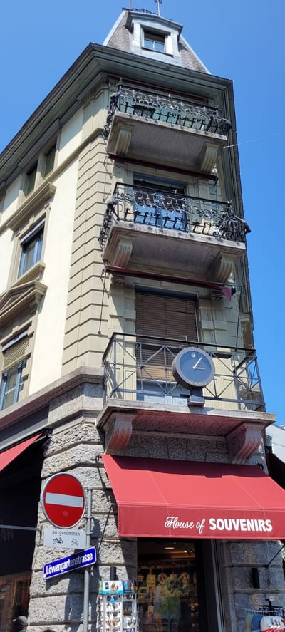 The image shows a street view in Wattens, Austria, focusing on the facade of a building. The building has a cream-colored exterior with stone detailing around the base and corners. It features multiple balconies with ornate black metal railings. A prominent dark clock with a white face and hands, displaying the time as approximately 12:08, is mounted on the railing of a mid-level balcony. The building has red awnings over storefronts at street level. One awning displays the text "House of SOUVENIRS" in white script. Below the awnings, there are signs, including a red circular "no entry" sign and a blue street sign that reads "Löwengartenstrasse". The sky above is clear and blue. The scene is captured from a low-angle perspective, emphasizing the height of the building. There are no people clearly visible or identifiable in the image.