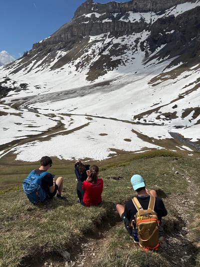 The image depicts a mountainous landscape in Mollens, Valais (VS), Switzerland, likely during a summer season due to the presence of grass and melting snow on the mountain. Three individuals are seated on a grassy slope, observing the mountain. One person wears a blue baseball cap and a hiking backpack, another is wearing a red shirt, and the third individual is slightly obscured. The backdrop includes a steep mountain face with patches of snow and rocky terrain. The sky is blue with minimal cloud cover, indicating a sunny day.