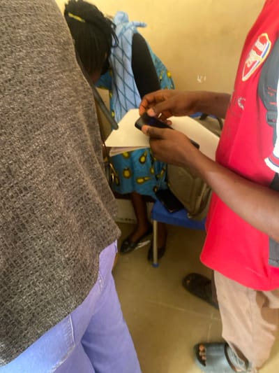 The image depicts an indoor scene in Baganakwo, Nigeria, involving three individuals. In the foreground on the left, an individual is partially visible, wearing a gray textured top and light purple trousers. To the right, another individual, wearing a red shirt with an Arsenal FC crest on the left chest and light brown trousers, is actively manipulating a dark smartphone with both hands over white sheets of paper. This individual wears dark open-toed sandals. In the background, a third individual is seated on a blue chair. This person wears a blue dress with a yellow circular pattern and a light blue patterned garment, and has dark braided hair with yellow accents. Black flat shoes are visible on their feet. A second dark smartphone rests on the blue chair beside the seated individual. The environment consists of plain, light-colored walls and a light brown floor.