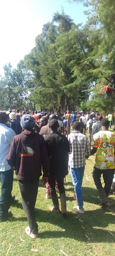 The image shows a group of people standing in a grassy clearing. The group consists of mostly men, though there are a few women visible in the background. Many are wearing casual clothing. The clearing is surrounded by trees, and the sky is clear. The image was taken in Isukha ICHINA, Kenya, at the location with the coordinates (latitude: 0.351, longitude: 34.781). The individuals in the foreground are walking toward the camera. One man in the foreground is wearing a black jacket with the word "Dreamwilder" printed on it in orange. Behind him, two men are wearing long pants, dark shirts, and boots. The man on the right is wearing a plaid shirt and blue pants. Another man in the foreground is wearing a yellow and green shirt with a geometric pattern. The people in the background appear to be standing and talking with each other. The light is bright, with no clouds in the sky.