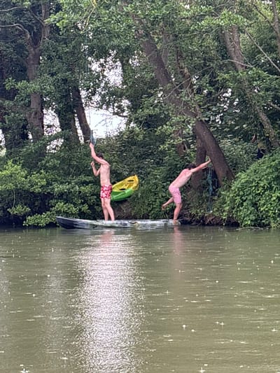 Two men in swimming trunks are about to jump into a body of water. One man is swinging from a tree branch above, wearing pink trunks, while the other is about to jump off a platform, wearing red floral trunks and carrying a yellow and green inflatable raft. Lush green trees and foliage surround the water's edge. The water appears to have a slightly reflective surface, and the image is rotated.