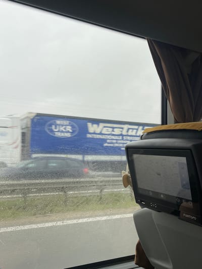 The image shows the view from the inside of a bus, looking out of the window. The window is covered in raindrops. A semi-trailer truck with the words "Westuk Internationale Spedition" can be seen through the raindrops. The truck is driving on a highway in the city of Osová Bítýška, Czechia, at the coordinates 49.325, 16.174. The truck is blue and has a white circle with the letters "UKR TRANS" written in it. The bus is moving forward and the truck is driving in the same direction. The image is taken from a passenger seat. There is a brown curtain in the upper right corner of the image. To the right of the window, there is a gray and black seatback with a screen attached to it. The screen shows a map with a red pin on it. It appears to be a navigation or tracking system. There is a small white fan mounted on the back of the seat.  The sky is overcast and the weather appears to be rainy.