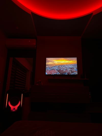 The image depicts an interior room dimly illuminated by red ambient lighting. A curved red LED strip light is recessed into the ceiling, which also features dark, angled panels. On the left wall, a white wall-mounted air conditioning unit is present. A large flat-screen television is mounted centrally on the far wall, displaying an aerial cityscape at sunset or sunrise, characterized by numerous red-tiled roofs, a body of water, and a long suspension bridge in the distance. Below the television, a dark, multi-drawer dresser or media console is visible, topped by a narrow shelf. A small, light-colored object, possibly a decorative skull or sculpture, rests on the shelf to the left of the television's vertical center. To the left of the dresser, an open doorway or archway leads to an adjacent space, revealing horizontal window blinds. In the lower-left foreground, a dark, vertical electronic device, possibly a speaker, features illuminated orange-red vertical light strips and a central red-lit emblem resembling stylized horns or a "U" shape. No individuals are present. The specific location within Erden, Bulgaria, is not visually identifiable from the image content.