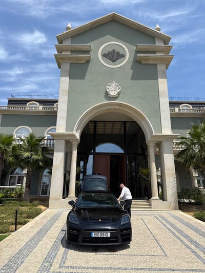 A medium, eye-level, outdoor shot, depicting a black car parked on a paved driveway in front of a building of classical architectural style, potentially located in Vila Verde, Portugal, based on the license plate. A man, dressed in a white shirt and black pants, leans into the open driver-side door of the car, with his back to the camera.
The building is painted in a light green hue, and features stone pillars and an arched entrance. A stone emblem is fixed above the entrance. Windows with semi-circular tops are visible on the second story. Palm trees are seen on either side of the entrance, suggesting a warm climate.
The driveway is paved with light-colored stones, arranged in a patterned design. The sky above is blue with scattered clouds.