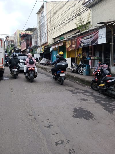 The scene depicts an overcast daytime view of a street in a commercial district, specifically described as being in Melakudikadu, India. However, prominent signage displays Indonesian language, such as "DIARUM SUPER," "TOKO TIGA SAUDARA," "PROMO CIKOR," and "ELITE."
The asphalt road, appearing somewhat worn with dark stains and patches, shows two visible lanes of traffic moving away from the viewer. In the immediate foreground/midground, three motorcycles are in motion. From left to right:
1. A dark-clad individual wearing a dark helmet rides a dark motorcycle, partially obscured by a white truck on the far left.
2. A motorcycle with two occupants. The rider wears a pink head covering (hijab), a light-colored top, and white pants, carrying a black backpack. A passenger is seated behind this rider. The license plate appears to begin with 'B 30XX FPU'.
3. A single rider wearing a yellow helmet, dark top, and dark pants on a dark motorcycle, which has a black storage box mounted on its rear. Its license plate is visible as 'B 3713 PII'.
A white car, possibly an SUV, is visible behind the middle motorcycle.
On the right side of the street, a raised concrete curb separates the roadway from a narrow sidewalk lined with a series of single to two-story commercial buildings. These buildings feature various shopfronts, some with partially open or closed metal roll-down shutters. Several motorcycles and scooters are parked along the curb. One notable parked scooter is black with yellow accents. A blue cylindrical trash bin is visible on the sidewalk. Overhead, numerous electrical and communication cables crisscross the sky. In the background on the left, a taller, light-colored building with a distinctive arched window structure stands prominently among other multi-story buildings.