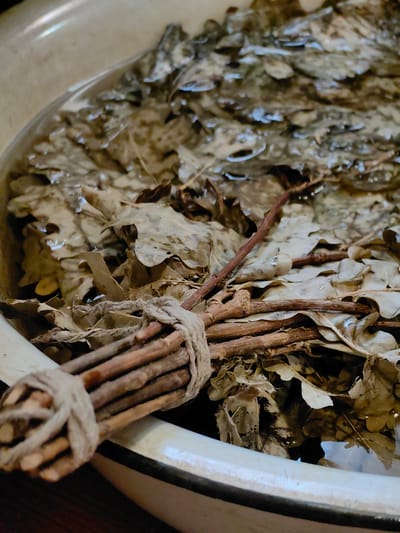 In the city of Fanipolski Sel'skiy Sovet, Belarus, a close-up shot captures a bundle of dried oak leaves submerged in water within a light-colored basin. The leaves, a dominant element, are brown and appear to be soaked, with some exhibiting signs of decay and damage. Interspersed among the leaves are thin, brown twigs, also submerged. A bundle of thicker twigs, bound together with rough, light-brown twine, is visible in the foreground, partially submerged and resting on the basin's rim and the leaves. The water's surface reflects the surrounding leaves and twigs, creating a slightly distorted view of the underwater elements. The basin's edge is visible in the lower left and right corners, and a dark surface, likely a table or floor, is visible beneath the basin. The lighting is soft, highlighting the textures of the leaves and twigs, and creating subtle reflections on the water.