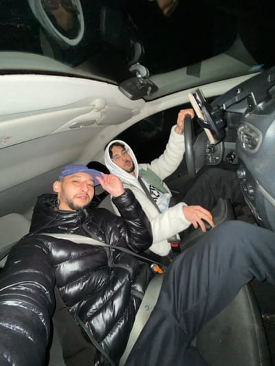 Two young men pose inside a car at night. One wears a shiny black puffer jacket and a blue baseball cap. The other is in a white hooded sweatshirt. The man in the sweatshirt holds the steering wheel. Another individual sits in the back seat behind the driver. The car's interior is visible with the dashboard and side mirror. Light reflects on the vehicle's surfaces.
