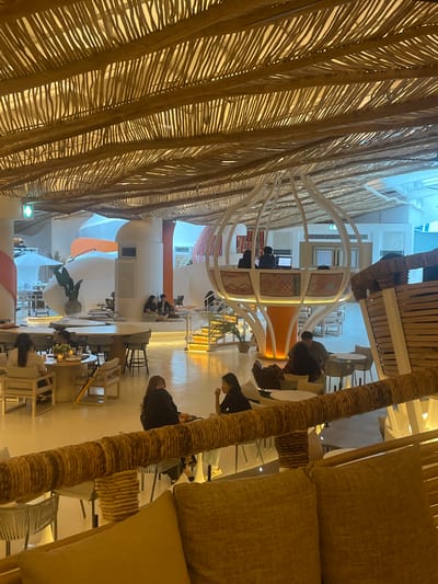 The image depicts the interior of a contemporary restaurant located in Gimpo-si, South Korea. The space features a distinctive ceiling constructed from woven bamboo-like material, casting warm shadows. The architectural design incorporates rounded, organic shapes with white and orange accents, creating a modern and airy ambiance. In the foreground, plush brown cushions are visible, suggesting a comfortable seating area.
Several patrons are seated at various tables and seating arrangements throughout the restaurant. In the mid-ground, a group of people are conversing at a round table with candles. Further back, individuals are seated in lounge areas and at small tables. A raised, pod-like structure houses what appears to be a DJ booth or service counter, with two individuals visible. A staircase with illuminated steps leads to this elevated area.
The floor is a light-colored, polished surface, reflecting the ambient light. The overall scene conveys a relaxed and social atmosphere, typical of a modern dining establishment. The presence of white umbrellas suggests an outdoor seating area or a connection to an outdoor space, though it is not explicitly shown.