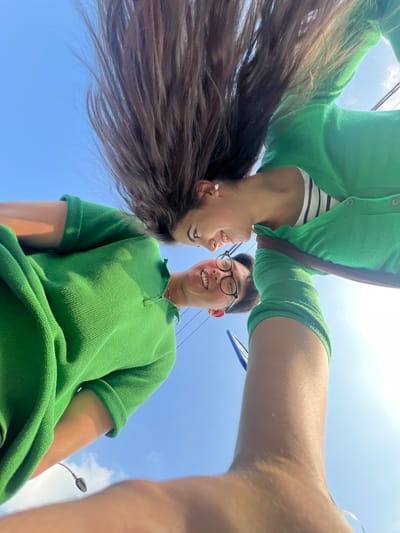 The media file is a low-angle, close-up shot of two people, an Asian male and a female with long brown hair, looking at each other and smiling. They are both wearing green tops. The female is positioned above the male, with her head upside down, and her hair cascades downwards. The male is looking up at her, his face filling the lower right portion of the frame. The background is a bright blue sky with some scattered white clouds, suggesting a clear, sunny day. The image appears to have been taken outdoors.
The subjects seem to be interacting playfully, with genuine smiles and eye contact, indicating a positive and possibly romantic relationship. The lighting suggests it is daytime, likely late morning or afternoon, given the brightness of the sky. There are no visible objects in the immediate foreground or background that provide specific location cues, other than the sky itself. The scene is casual and intimate, possibly a selfie taken by one of the individuals. No text is visible in the image.