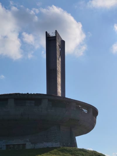 The image depicts the Buzludzha Monument, a Brutalist architectural relic located in Shipka, Bulgaria. The monument, a massive concrete structure resembling a flying saucer, is situated atop a grassy hill under a bright blue sky dotted with scattered white clouds. Sunlight casts a clear light on the scene, suggesting it is daytime, likely mid-morning to late afternoon.
The primary subject is the imposing monument itself. A tall, angular tower rises from the center of the circular main building, topped with a small platform featuring metal fencing. A star, a symbol often associated with socialist regimes, is visible on the side of the tower. The main structure is characterized by its wide, curved base, which shows signs of decay and weathering, including broken sections and visible rebar. Grassy slopes lead up to the monument, some of which are illuminated by the sun.
Although no people are explicitly visible, the presence of scaffolding on the tower suggests potential ongoing maintenance or restoration work, or perhaps that it is a site visited by people. The monument, abandoned and in disrepair, evokes a sense of historical significance and perhaps melancholy due to its state of decay, contrasting with the vibrancy of the sky.
Graffiti is visible on the lower, more accessible parts of the monument's exterior walls, adding another layer to its current state. Some text, likely inscriptions or graffiti, is discernible on the curved lower section of the monument, though it is difficult to read clearly due to the distance and the angle of the photograph. The overall impression is one of a monumental structure from a past era, now standing as a silent testament to history against a backdrop of a clear, serene sky.