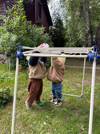 Two children interact with a white metal clothes drying rack outdoors on a grassy lawn in Tomilino, Russia. One child, wearing brown corduroy pants and a tan fleece jacket, holds a soccer ball. The other child wears blue jeans and a similar fleece jacket. In the background, a dark brown wooden house is visible, surrounded by lush greenery. The sky is overcast.